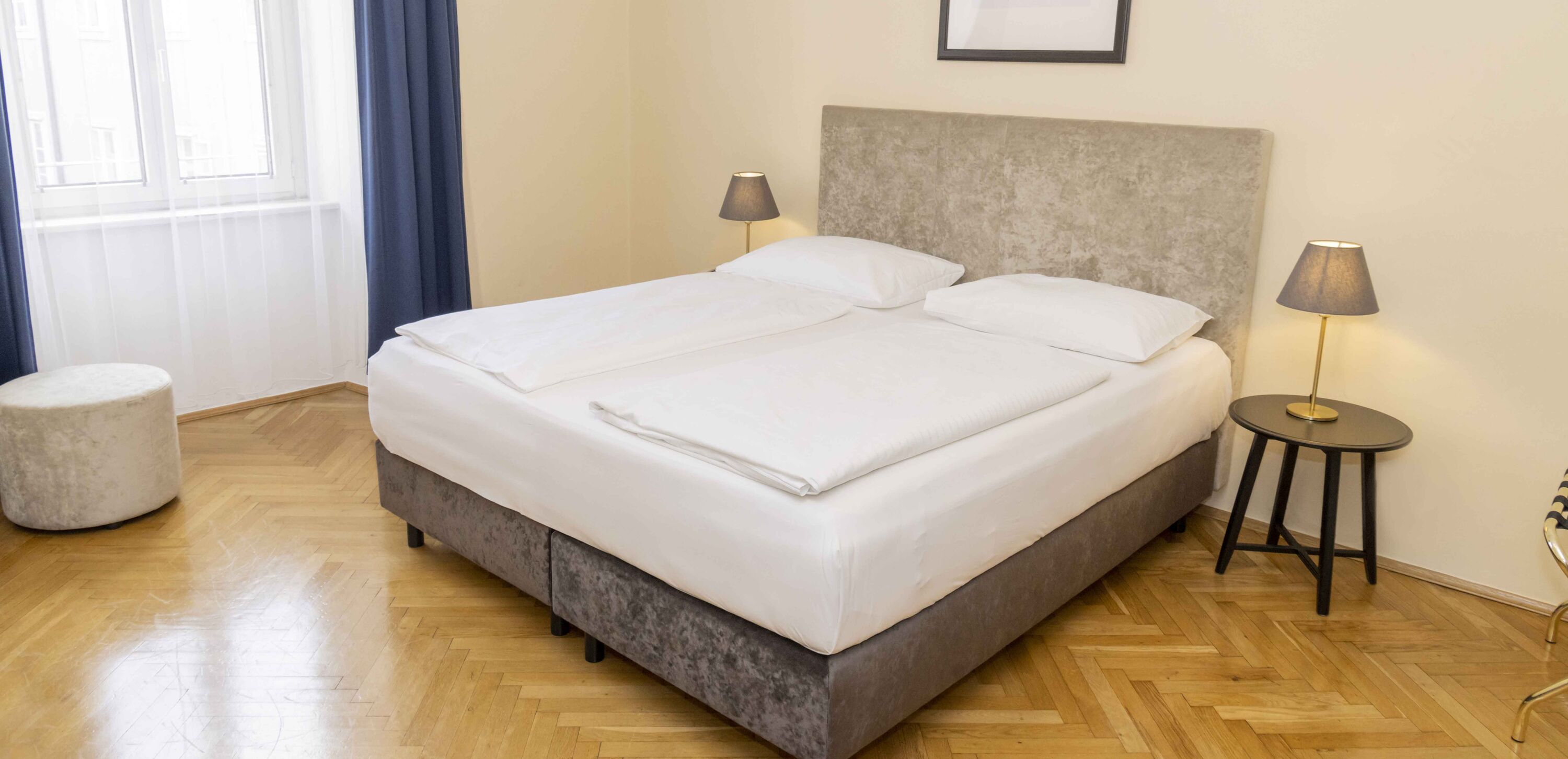 Foto zeigt großzügiges Zweibettzimmer mit modernem grauen Doppelbett, Samt-Pouf und Blick zum Fenster.