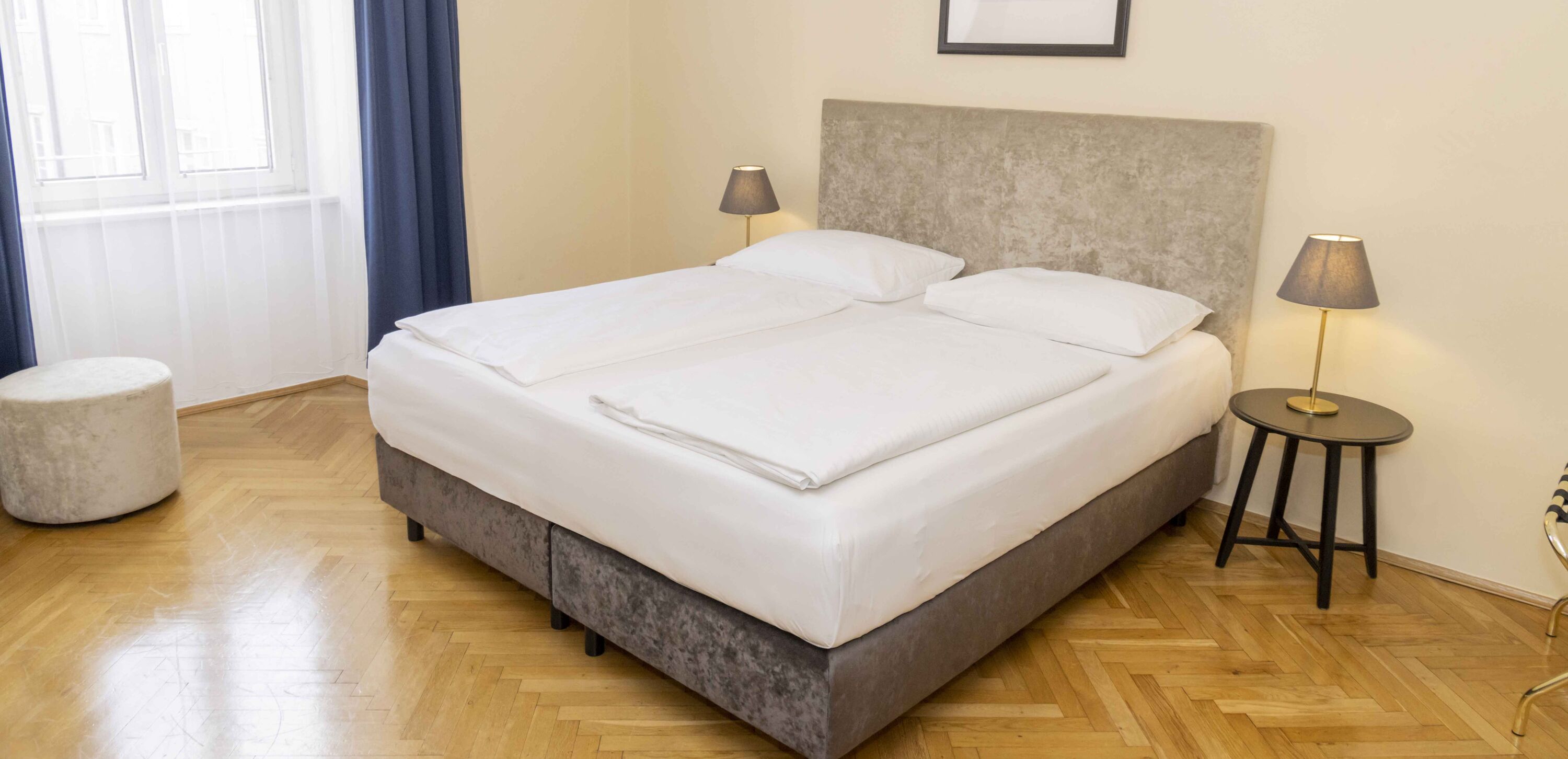 Foto zeigt großzügiges Zweibettzimmer mit modernem grauen Doppelbett, Samt-Pouf und Blick zum Fenster.
