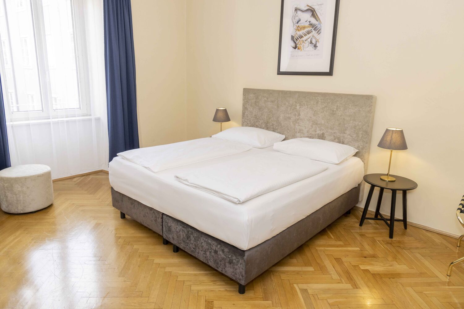 Foto zeigt großzügiges Zweibettzimmer mit modernem grauen Doppelbett, Samt-Pouf und Blick zum Fenster.