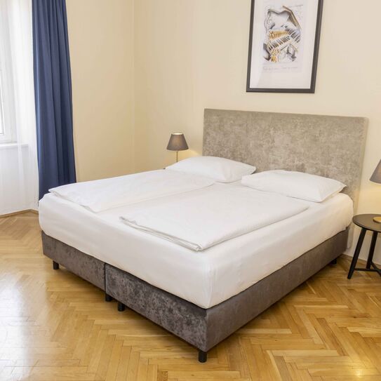 Foto zeigt großzügiges Zweibettzimmer mit modernem grauen Doppelbett, Samt-Pouf und Blick zum Fenster.