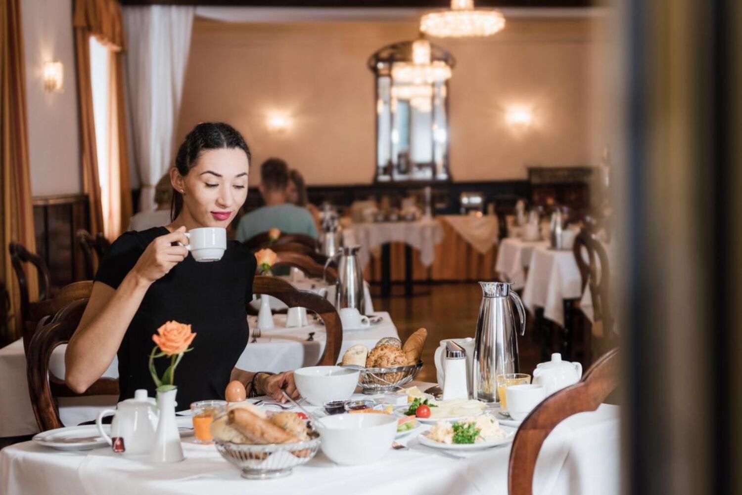 Foto zeigt Frau mit dunklen Haaren an einem Frühstückstisch sitzend und Kaffee trinkend.