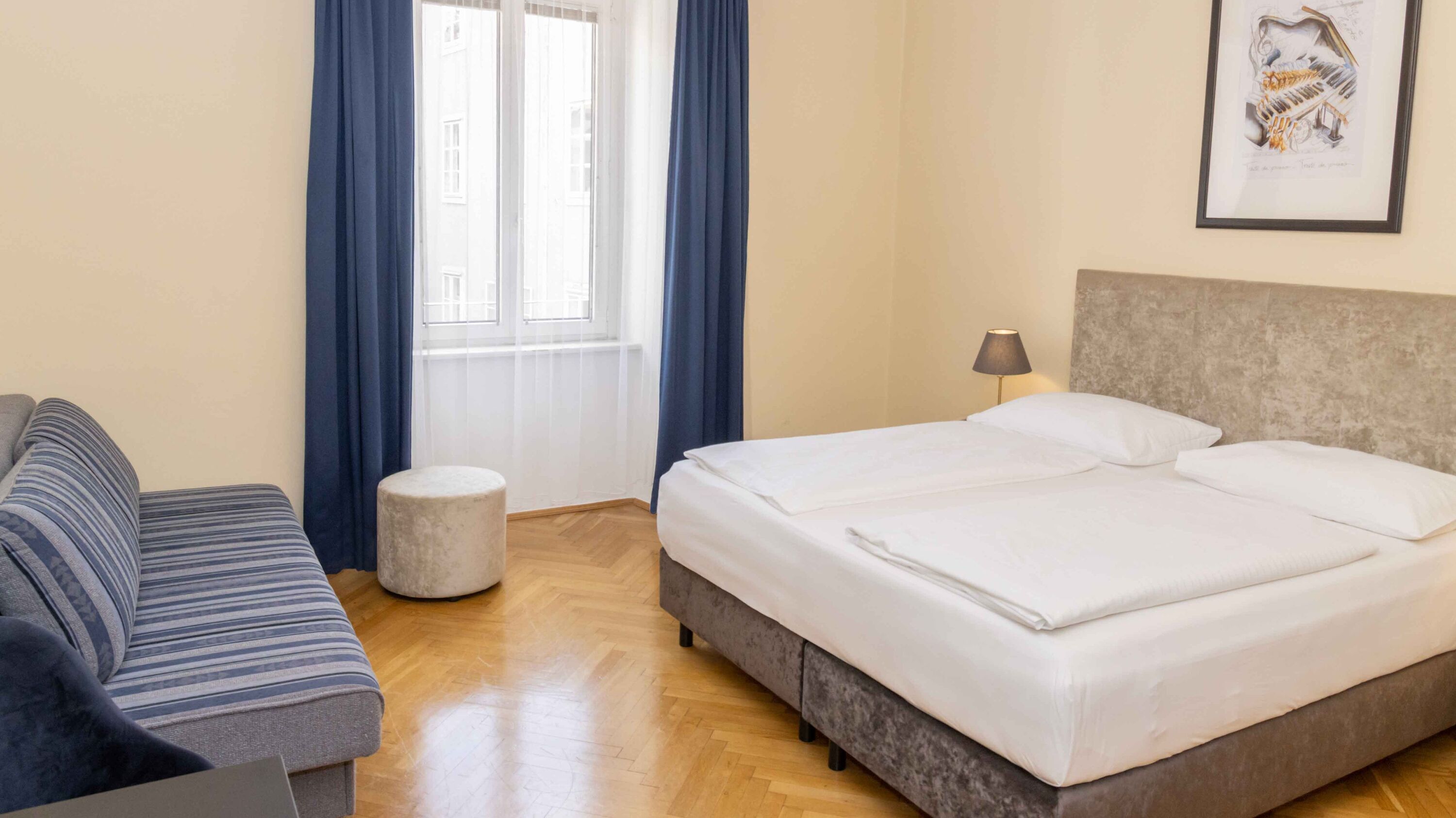 Foto zeigt großzügiges Dreibettzimmer mit modernem Doppelbett, blauer Couch und Blick zum Fenster.