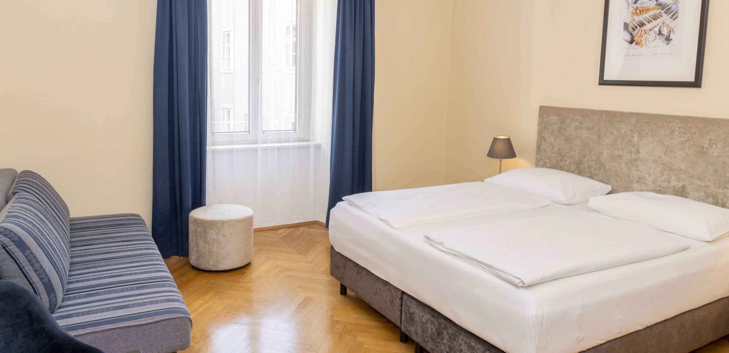 Foto zeigt großzügiges Dreibettzimmer mit modernem Doppelbett, blauer Couch und Blick zum Fenster.